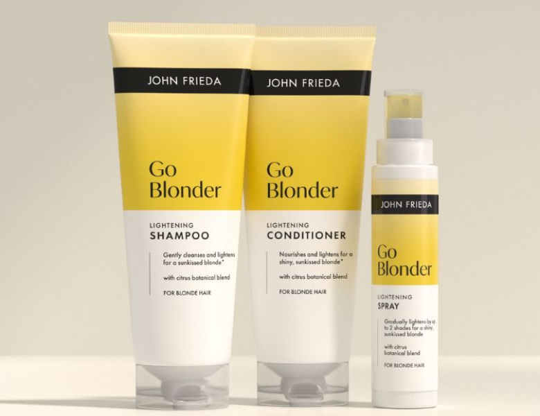 Her ses et billede med hårplejeprodukter fra John Frieda placeret mod en lys baggrund. På produkterne står teksten “Go Blonder”, og serien indeholder shampoo, conditioner og spray. Nederst står teksten “BLONDE”.