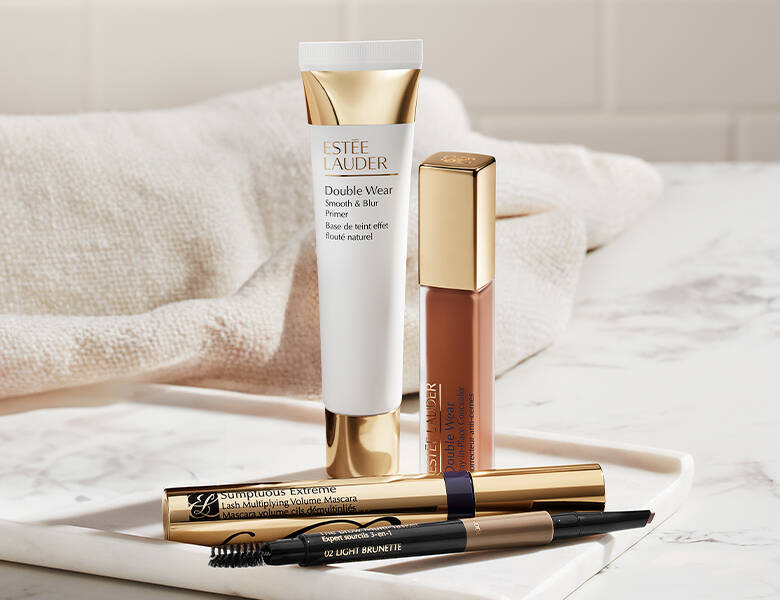 Her ses fire makeupprodukter fra Estée Lauder som ligger på et lyst bord; en primer, en lipgloss, en mascara og en brynpen.