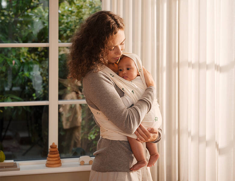 En mor holder sit barn tæt ind på kroppen og sen ned på det. Barnet er i en hvid bæresele eller strækvikle. Lyst rum med udsigt til grøn have gennem vindue bagved.