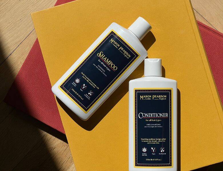 Her ses to hvide Mason Pearson produkter med sorte labels. De ligger på en gul bog med en rød bog derunder. 