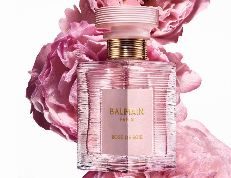 Her ses et billede af en parfumeflakon fra Balmain placeret foran store lyserøde blomster mod en lys baggrund. På flakonen står teksten “BALMAIN PARIS ROSE DE SOIE”. Flakonen er i lyserød nuance med guldfarvede detaljer.