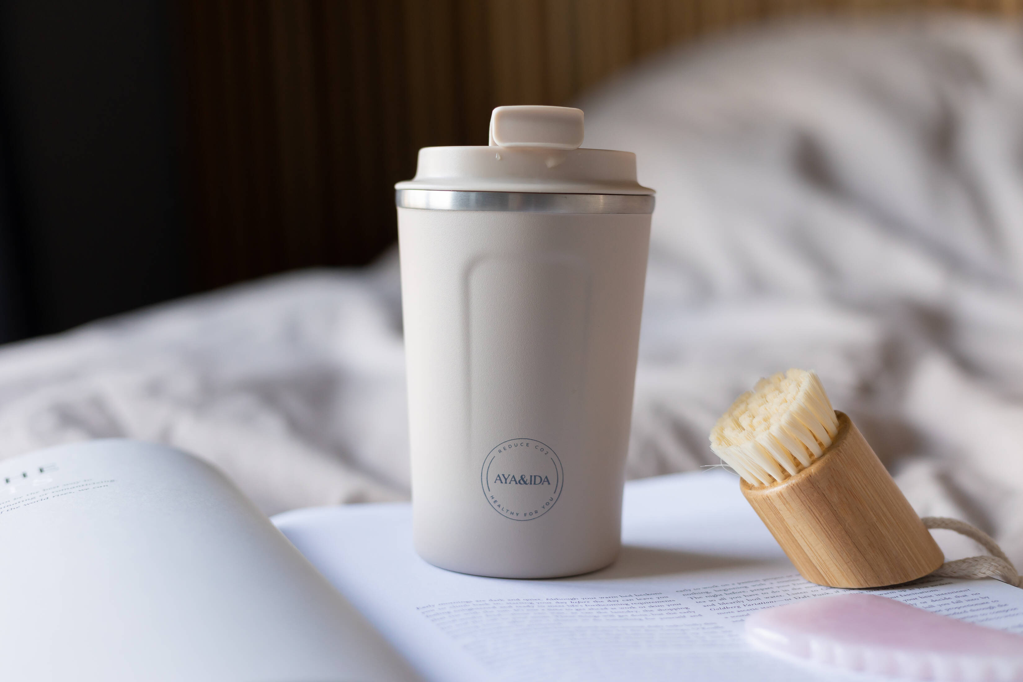 Her ses en beige to-go-kop, som står på en åben hvid bog, som ligger i en neutralfarvet seng. 
