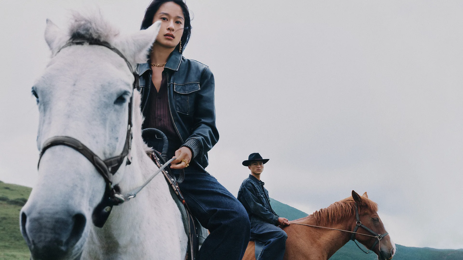 Two models riding horses across an open landscape under an overcast sky, creating a cinematic and atmospheric 