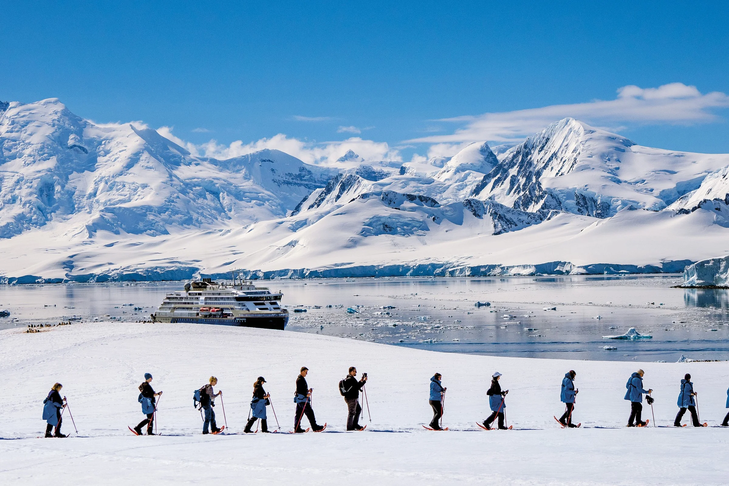 A group of people in winter gear walk in a line across snowy terrain near a calm icy bay, with a large cruise ship and snow-covered mountains in the background under a clear blue sky.