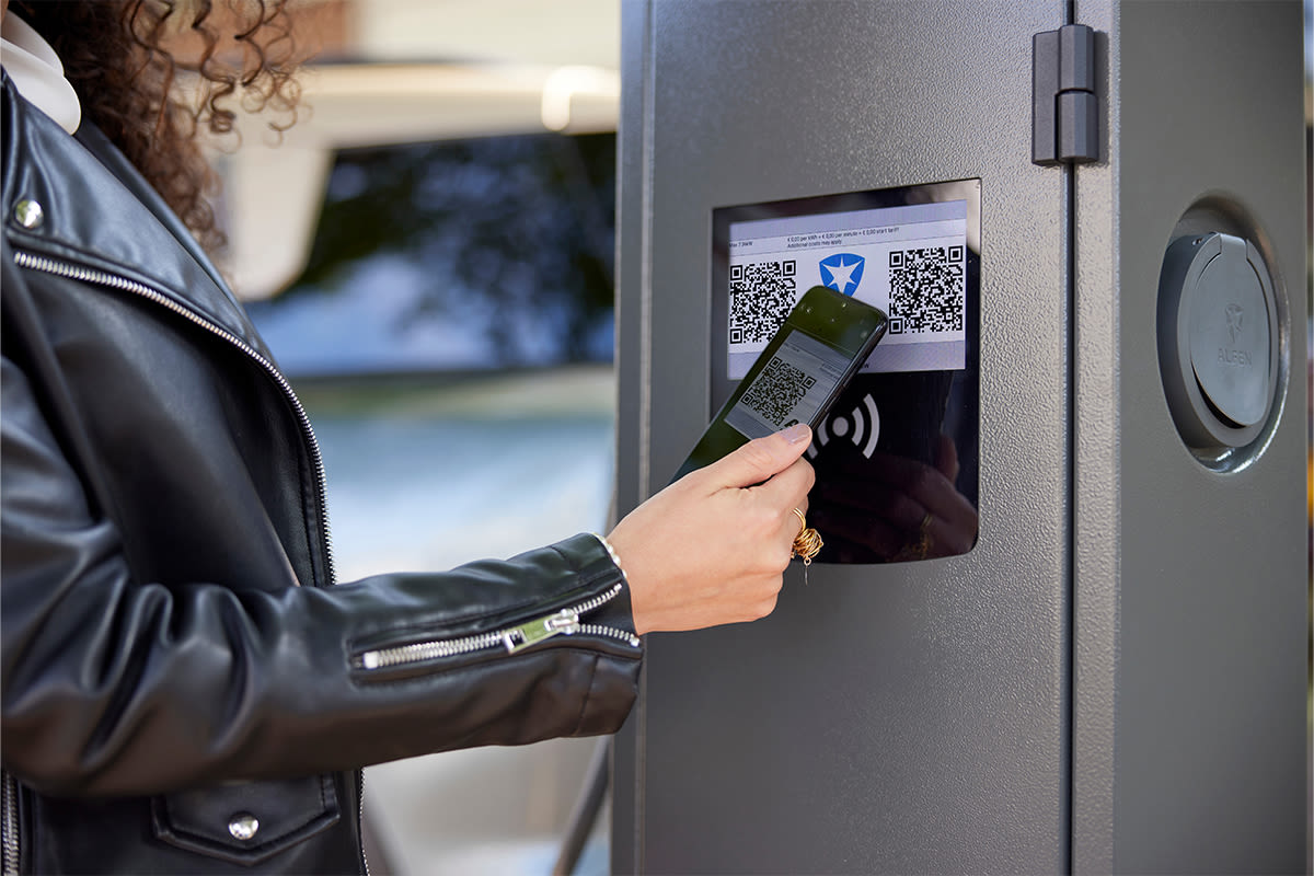 Een vrouw scant met zijn telefoon een QR-code op een laadstation. Ze draagt een zwart leren jack, wat een gevoel van moderne technologische interactie oproept.