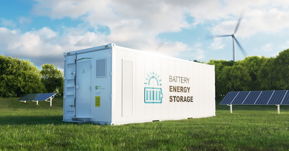 Witte batterijopslagcontainer op een groen veld met zonnepanelen en een windturbine op de achtergrond, onder een blauwe lucht met wolken.
