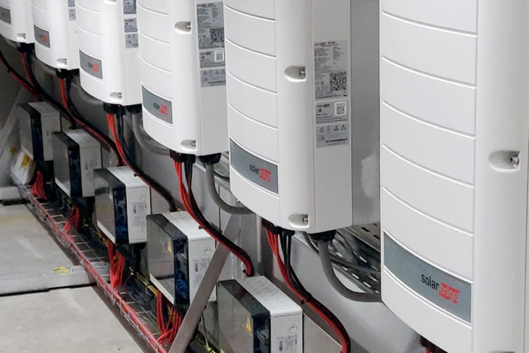 Een rij witte SolarEdge power optimizers, gemonteerd op een muur, met rode en zwarte kabels die eronder lopen. De opstelling ziet er georganiseerd en technisch uit.