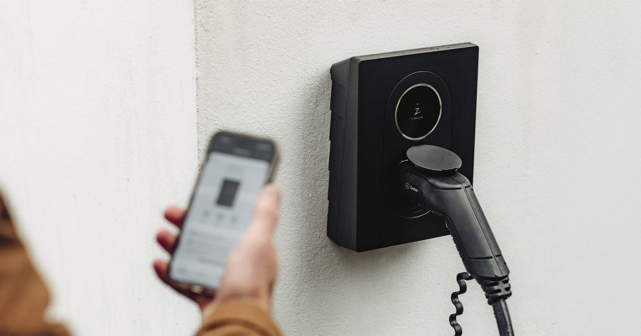 Eine Person hält ein Smartphone in der Nähe der an einer weißen Wand montierten Zaptec Go 2 Ladestation.