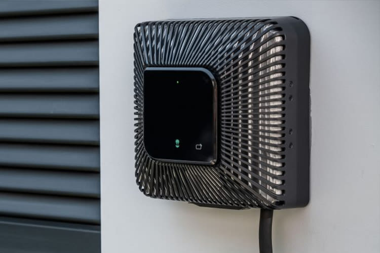 Aan de muur gemonteerde laadpaal voor elektrische voertuigen met een strak zwart, geribd ontwerp en een aangesloten kabel, tegen een witte muur, wat moderniteit uitstraalt.