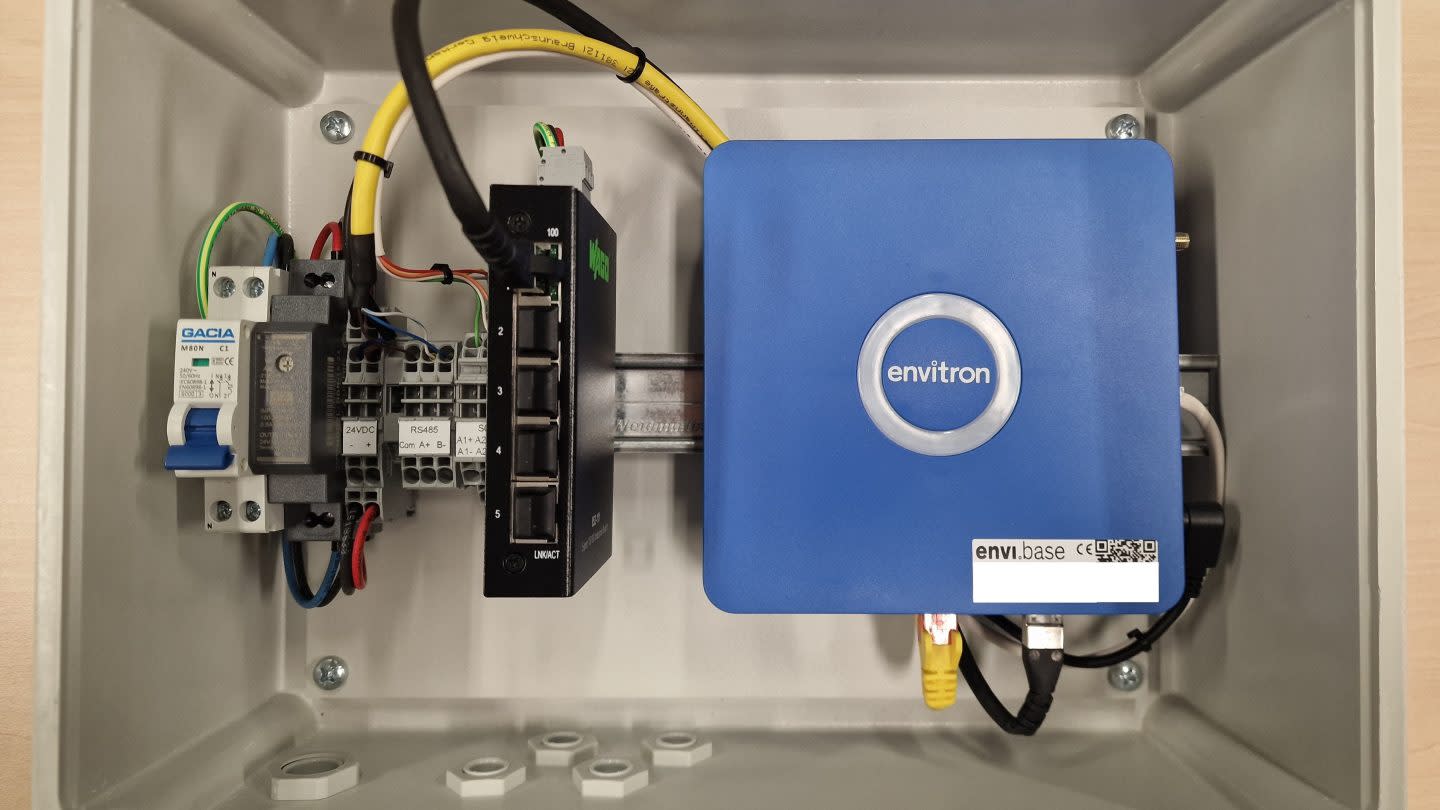 Interieur van een elektrisch paneel met rechts een blauw Envitron apparaat en verschillende aangesloten kabels. Neutrale tinten, technische opstelling.