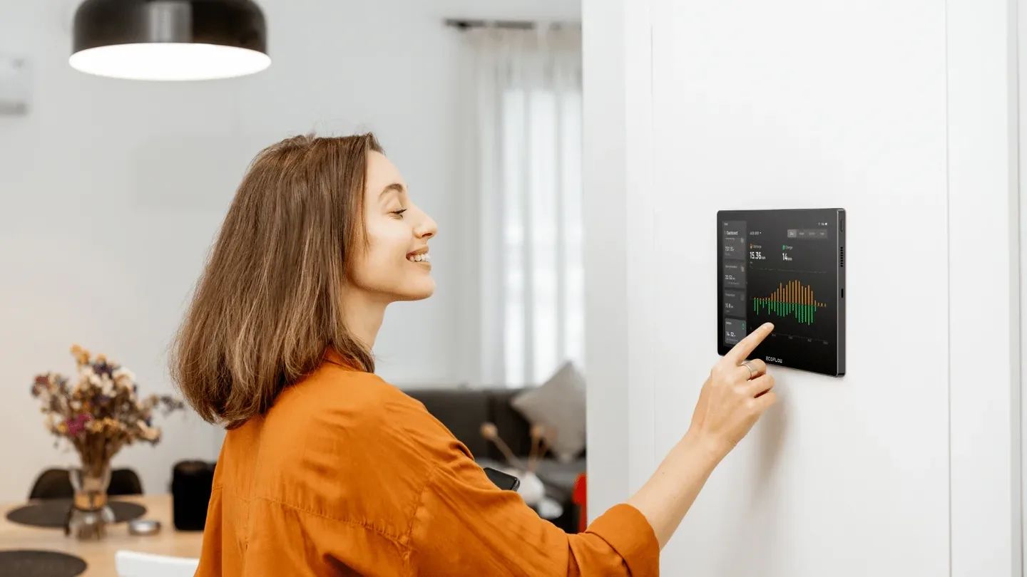 Een vrouw in een oranje shirt bedient een aan de muur gemonteerd “EcoFlow PowerInsight” smart home apparaat waarop kleurrijke grafieken worden weergegeven. De ruimte is modern en goed verlicht en heeft een warme, gezellige sfeer.