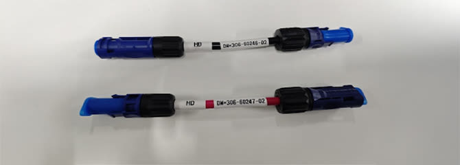 Zwei kurze Kabel mit blauen und schwarzen Steckverbindern an beiden Enden liegen parallel auf einer weißen Oberfläche. Jedes Kabel ist mit alphanumerischen Codes gekennzeichnet.