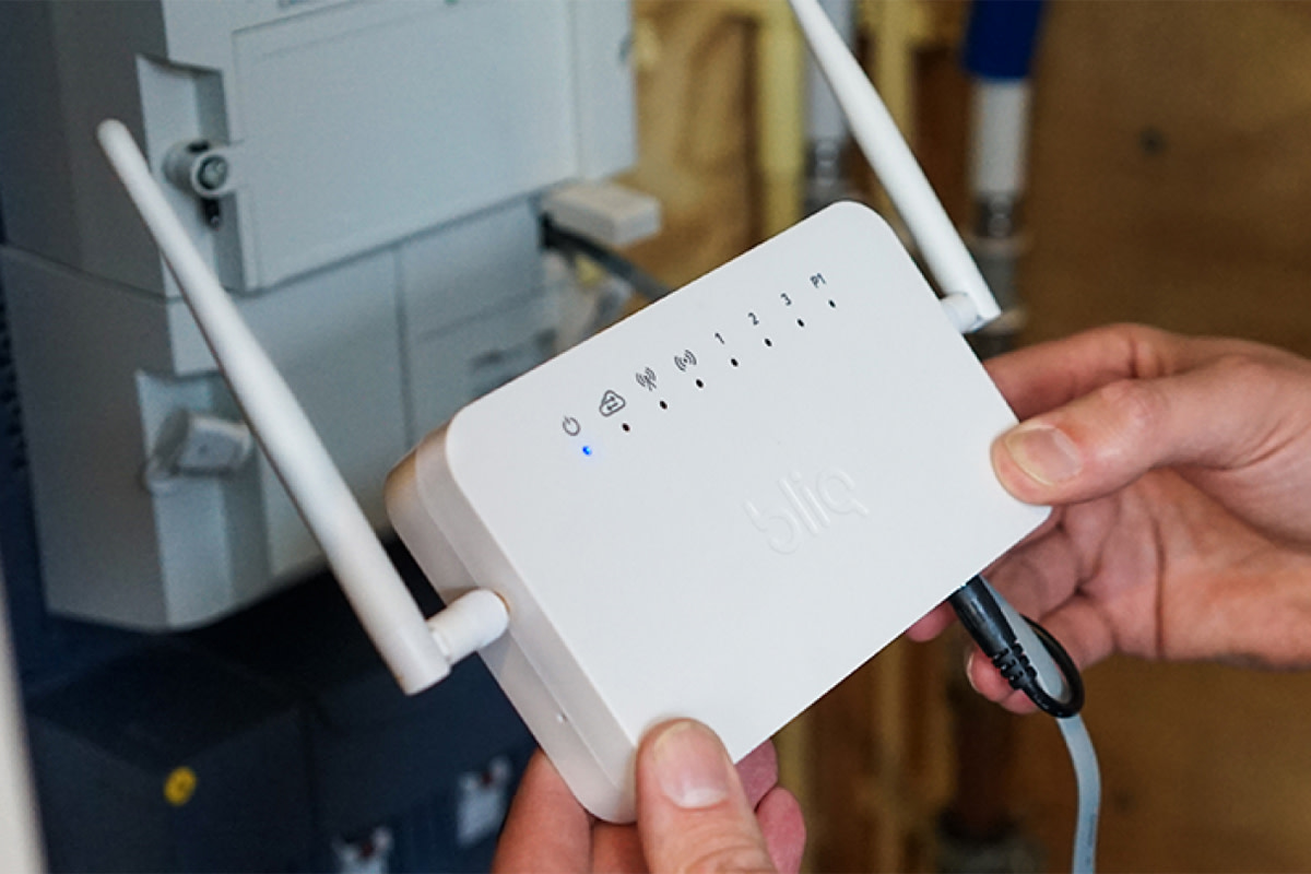 Een persoon houdt een witte router vast met de naam Bliq EMS, voorzien van twee antennes en indicatielampjes. De router bevindt zich bij een muur met elektronische apparatuur, wat wijst op installatie of configuratie.