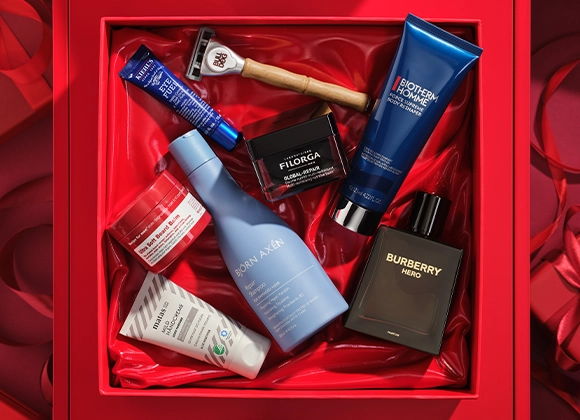 Bilde som viser flere skjønnhetsprodukter arrangert i en rød gaveeske. Produktene inkluderer en blå tube med Kiehl's Eye Fuel, en barberhøvel fra Bulldog, en svart boks med Filorga Global-Repair, en blå tube med Biotherm Homme Force Supreme Body Reshaper, en svart flaske med Burberry Hero parfyme, en blå flaske med Björn Axén Repair Shampoo, en hvit tube med Matas Men Ansiktskrem, og en rød boks med Recipe for Men Ultra Soft Beard Balm. Bakgrunnen er rød.
