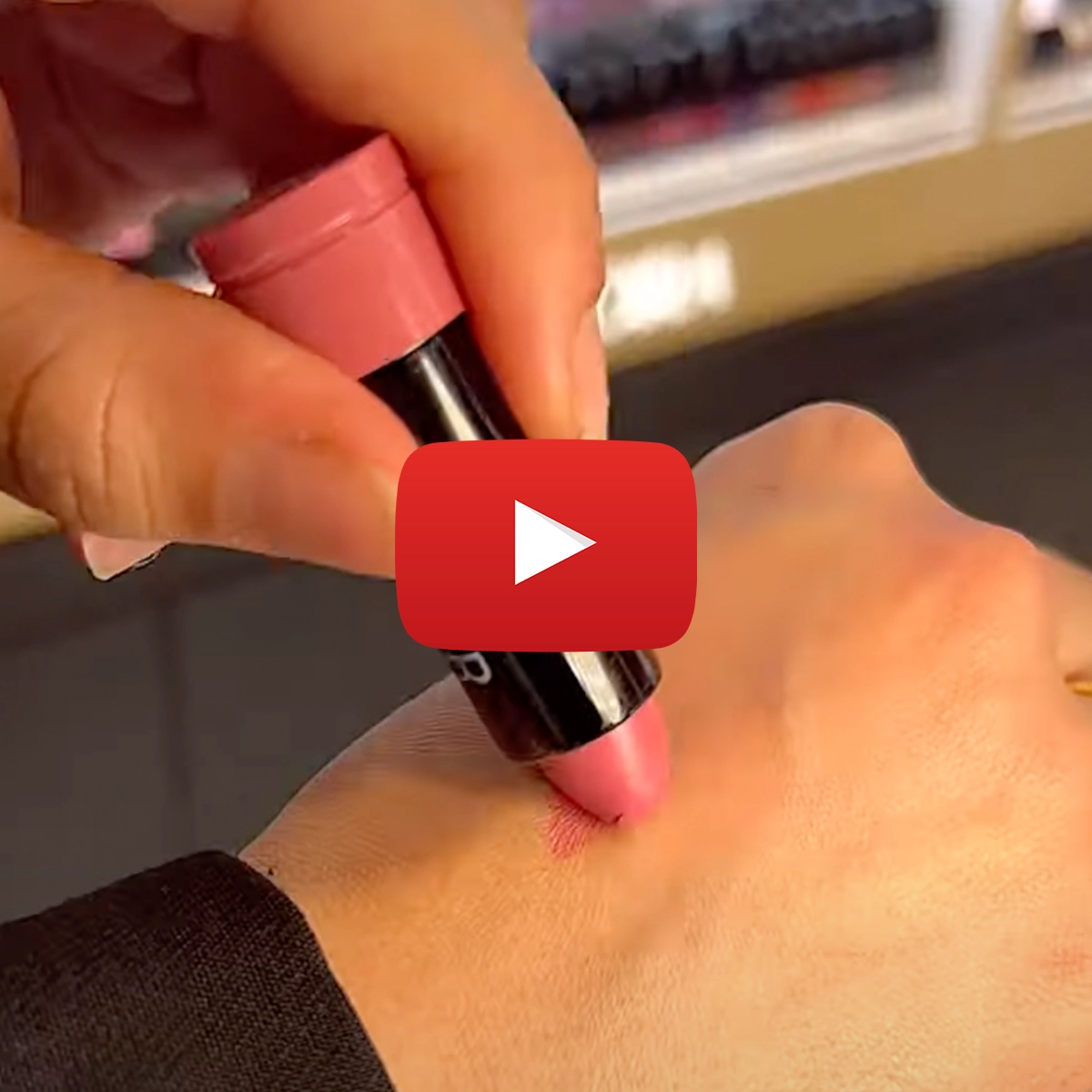 En hånd påfører en rosa leppestift på en annen hånd for å vise fargen. Et Youtube play-tegn er plassert midt på bildet.