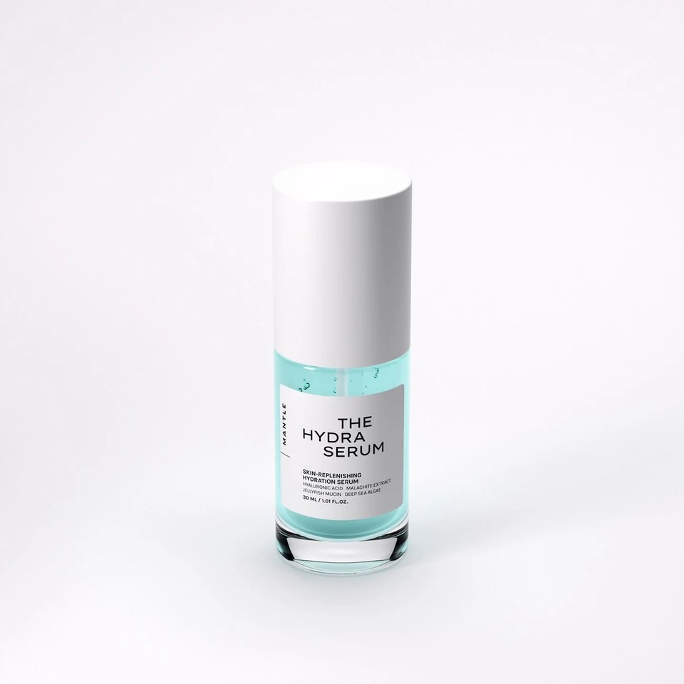 Hudvårdsserum i glasflaska med turkos vätska och vit kork mot ljus bakgrund, med etiketten "THE HYDRA SERUM" och text som "Skin-Replenishing Hydration Serum", "Hyaluronic Acid + Malachite Extract + Irish Moss + Deep Sea Algae" samt "30 ml / 1.01 fl. oz.".