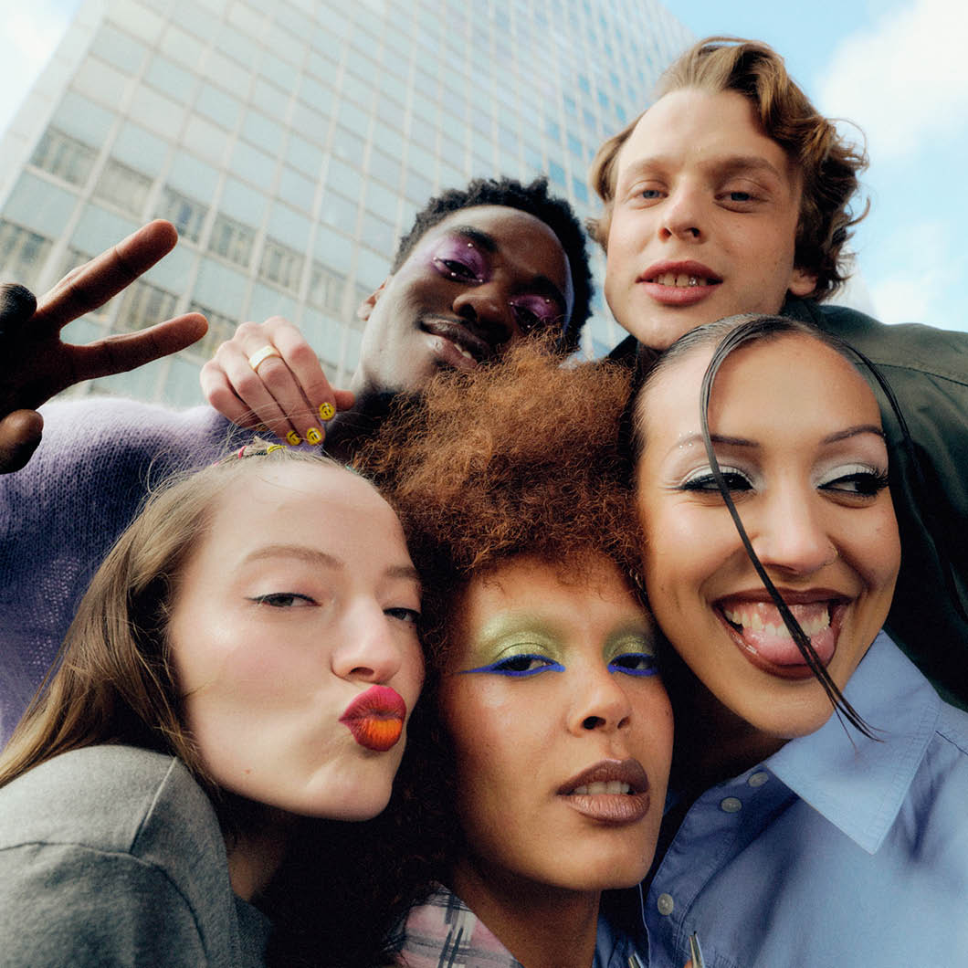 Fem unge personer tar en nærbilde-selfie utendørs, ler og lager lekne ansiktsuttrykk. De har fargerik sminke og står tett sammen med bygninger og himmel i bakgrunnen.