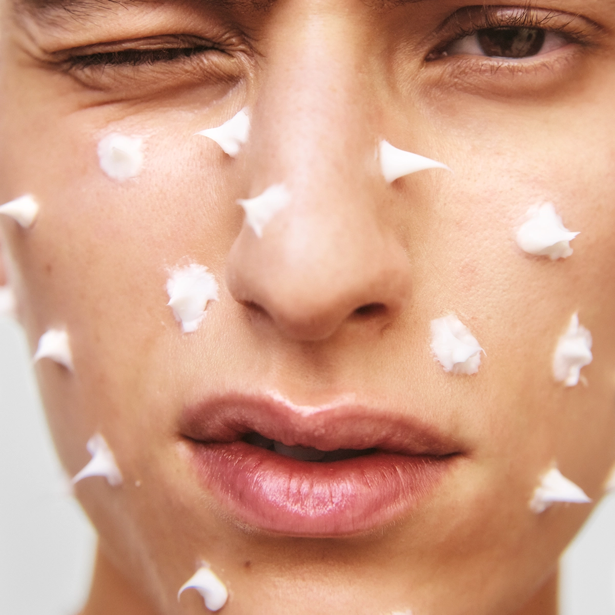 Lähikuva kasvoista, joille on levitetty pieniä valkoisia kasvovoidepisteitä tasaisesti poskille ja suun ympärille. Painotus ihonhoitoon ja kosteuttavaan kasvovoiteeseen ennen levitystä.