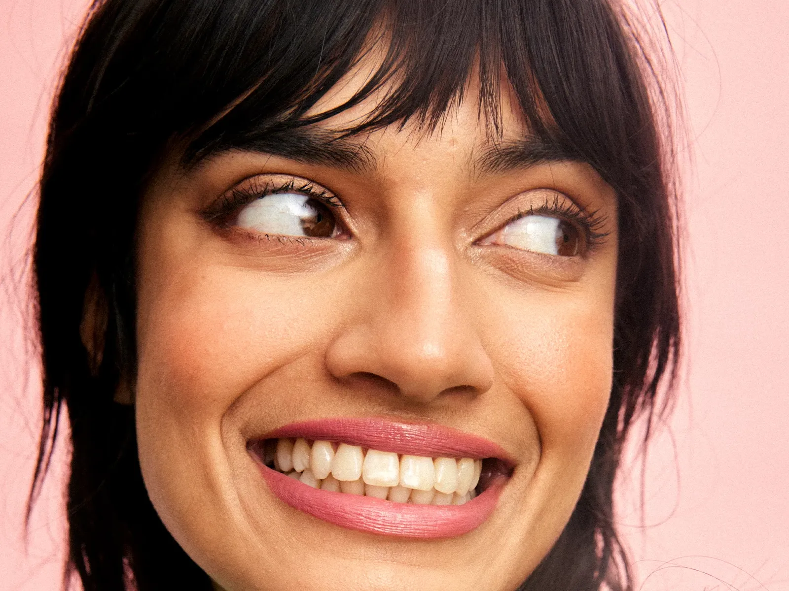 Nærbilde av en smilende person med mørkt hår og rosa leppestift mot en rosa bakgrunn.