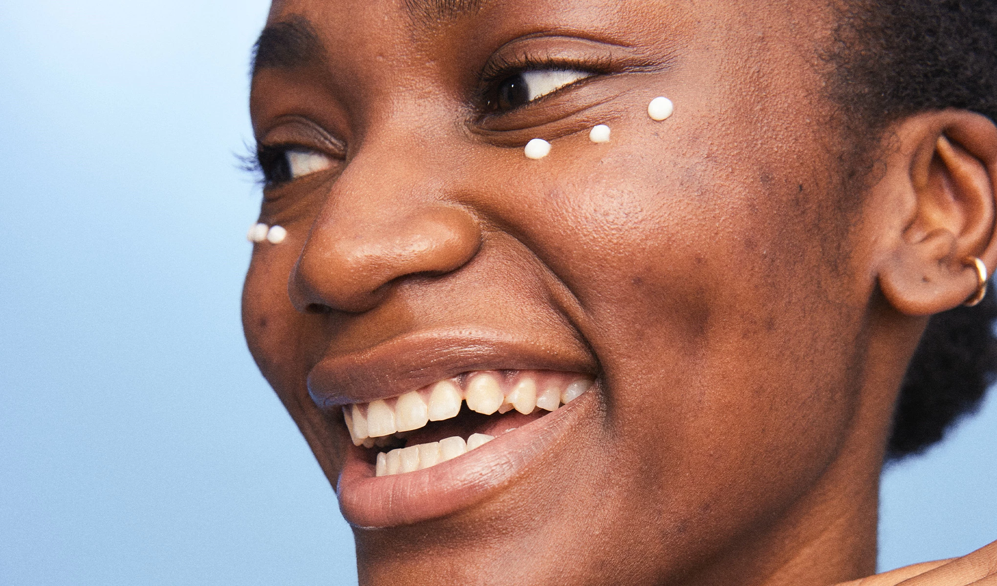 Hymyilevä nainen sinistä taustaa vasten, pienet pisarat silmänympärysvoidetta silmiensä alla. 