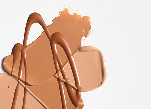 Nærbilde av kremet foundation eller concealer i beige nyanse smurt ut i brede strøk med blanke dråper oppå mot lys bakgrunn. Flytende makeup i nøytral tone med synlig tekstur og dekkende finish.