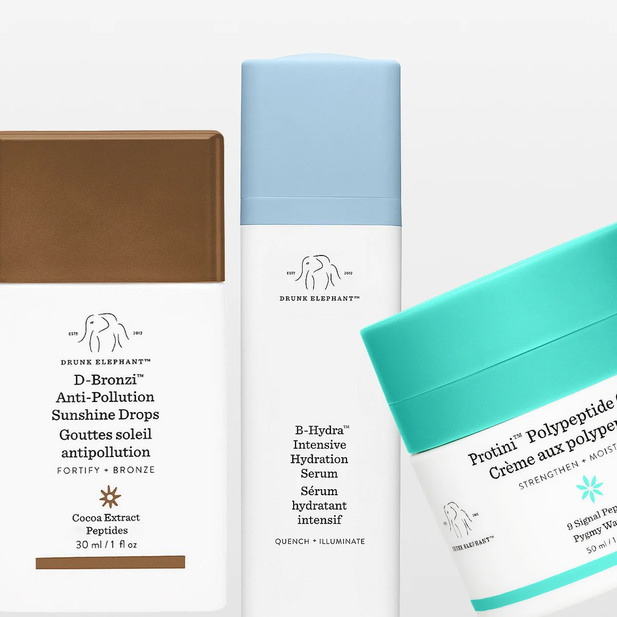 Tre hudpleieprodukter fra Drunk Elephant vises mot lys bakgrunn: D-Bronzi Anti-Pollution Sunshine Drops i brun flaske, E-Hydra Intensive Hydration Serum i hvit og blå emballasje og Protini Polypeptide Cream i en krukke med turkist lokk.