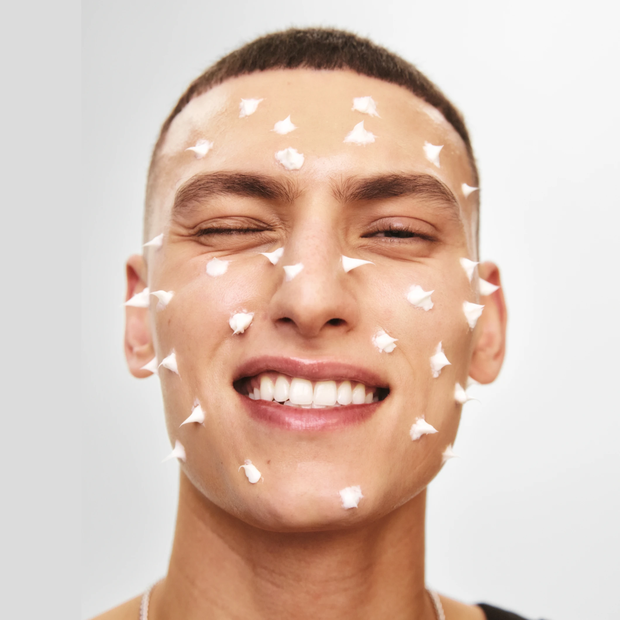 Lähikuvassa henkilö, jolla on lyhyet hiukset ja valkoista kasvovoidetta otsalla, poskilla ja leuassa. Henkilö hymyilee vaaleaa taustaa vasten, ja kuvassa korostuvat ihonhoito ja kosteuttavat skincare-tuotteet.