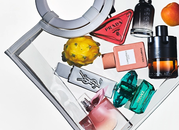 Flere parfymeflasker arrangert i en gjennomsiktig håndveske sammen med frukt. Synlige parfymer fra Prada, BOSS, Yves Saint Laurent, Dolce & Gabbana, Versace og Lancôme i ulike fargede glassflasker.