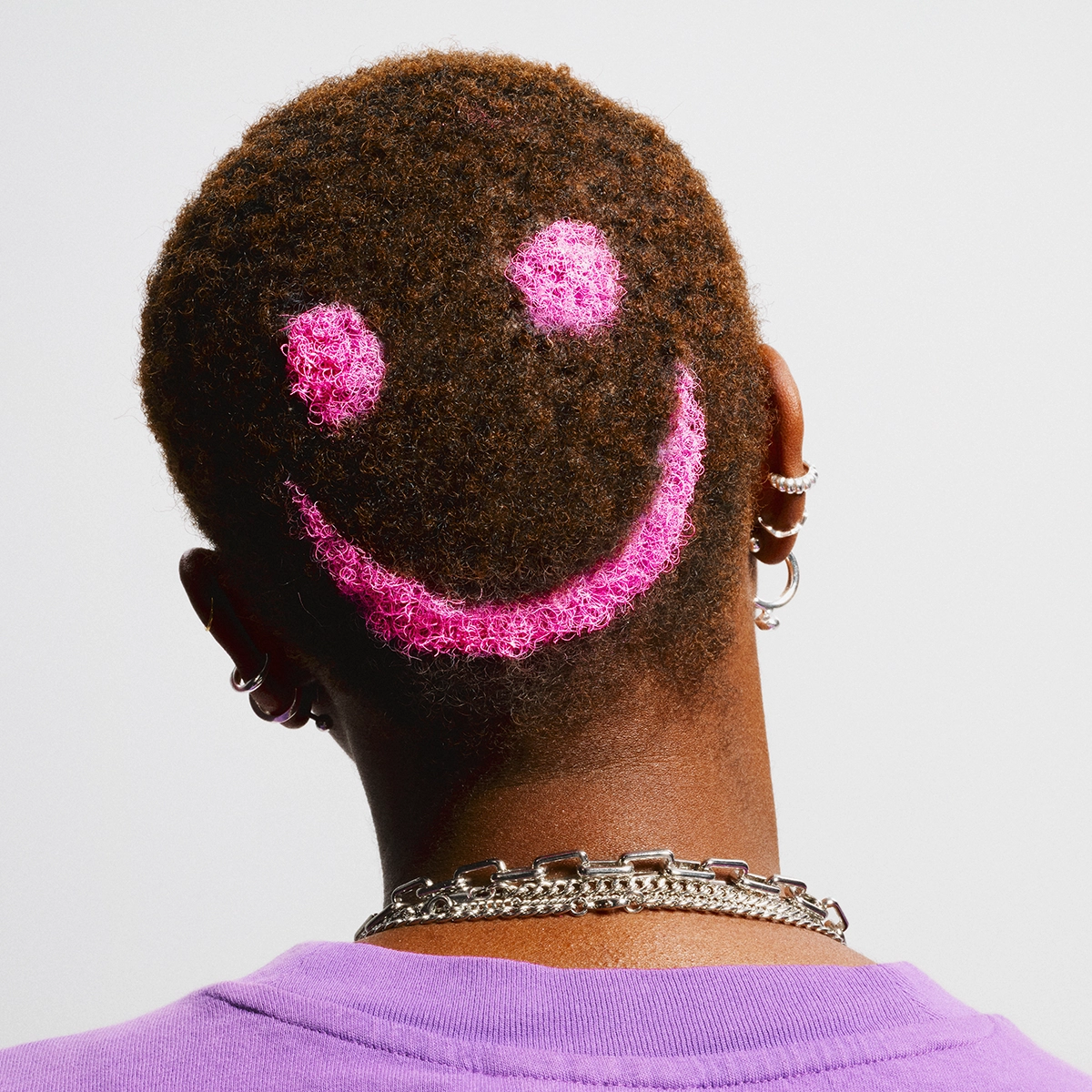 Bakhuvud med kort, lockigt hår där ett rosa smiley-motiv är infärgat i håret. Personen bär lila tröja, flera örhängen och ett silverfärgat halsband.