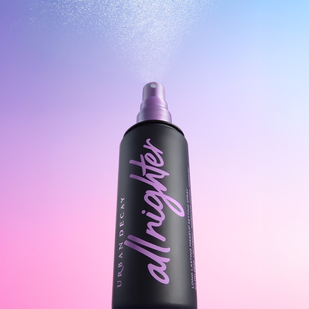 En svart sprayflaske med lilla tekst "all nighter" og "URBAN DECAY" sprayer tåke mot en gradient bakgrunn i rosa og blått.