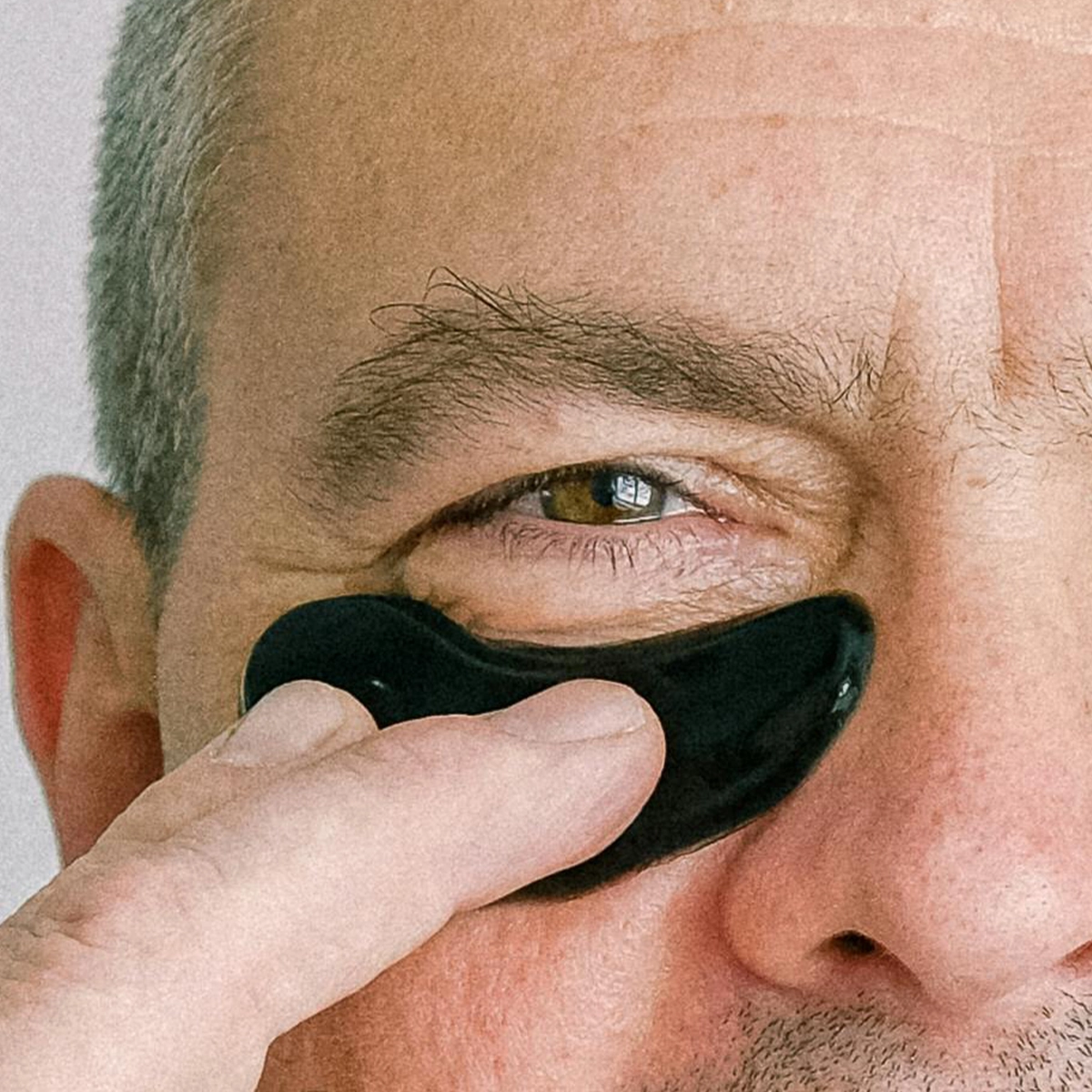Närbild på en person med en svart ögonmask under ögat som han håller fast med ett finger. Personen är gråhårig och bakgrunden är ljusgrå.
