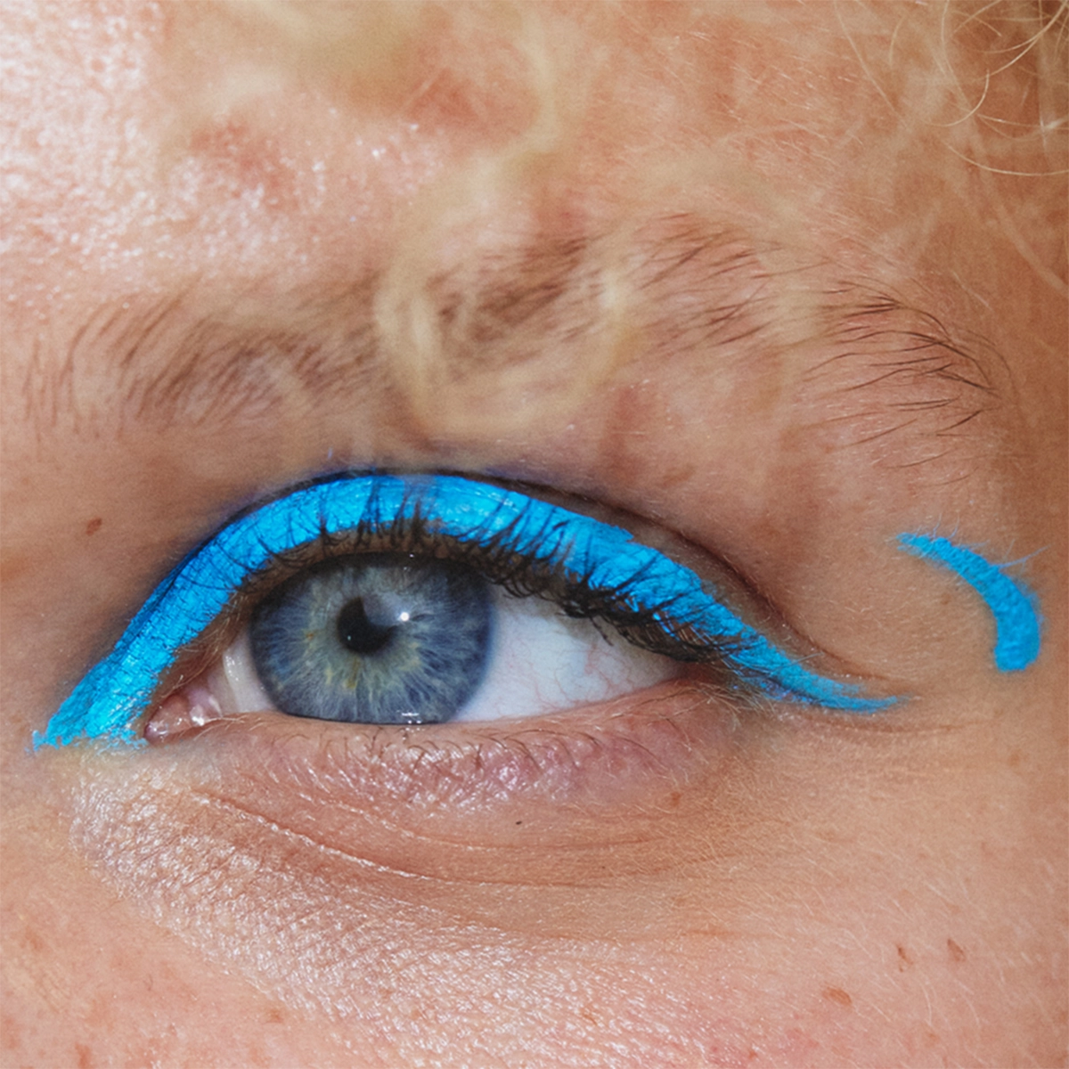 Nærbilde av øyne med intens turkisblå eyeliner langs de øvre øyelokkene, trukket ut i grafiske vinger ved øyekrokene. Små turkisblå detaljer er synlige utenfor begge øynene.