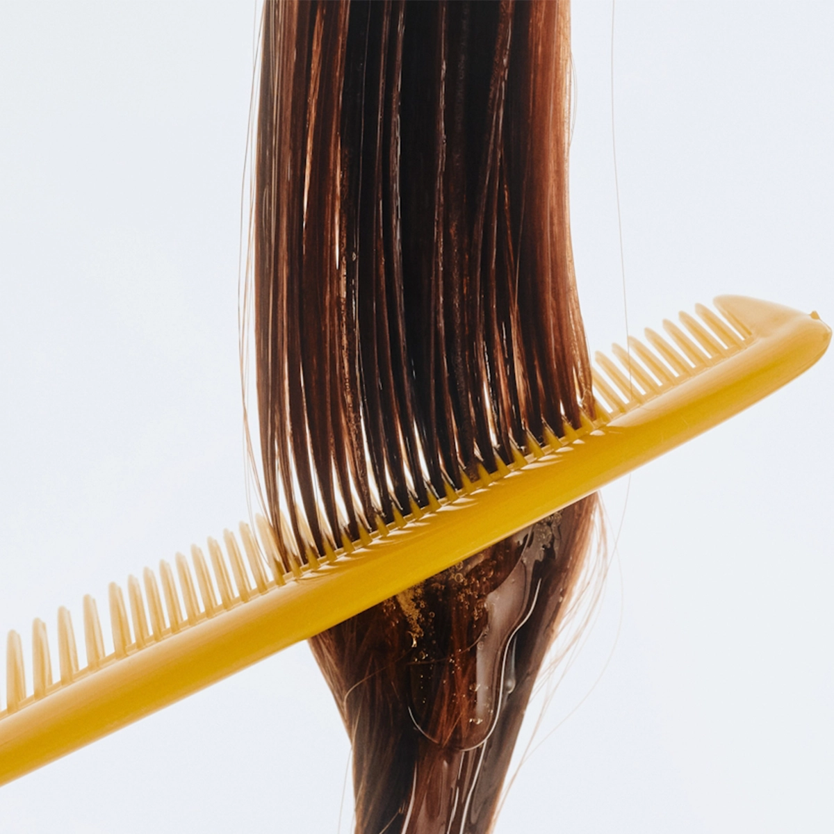 Närbild på en gul kam som dras genom långt, blött brunt hår med synlig hårinpackning eller balsam som fördelas jämnt i längderna. Fokus på hårvård och utredning mot ljus bakgrund.