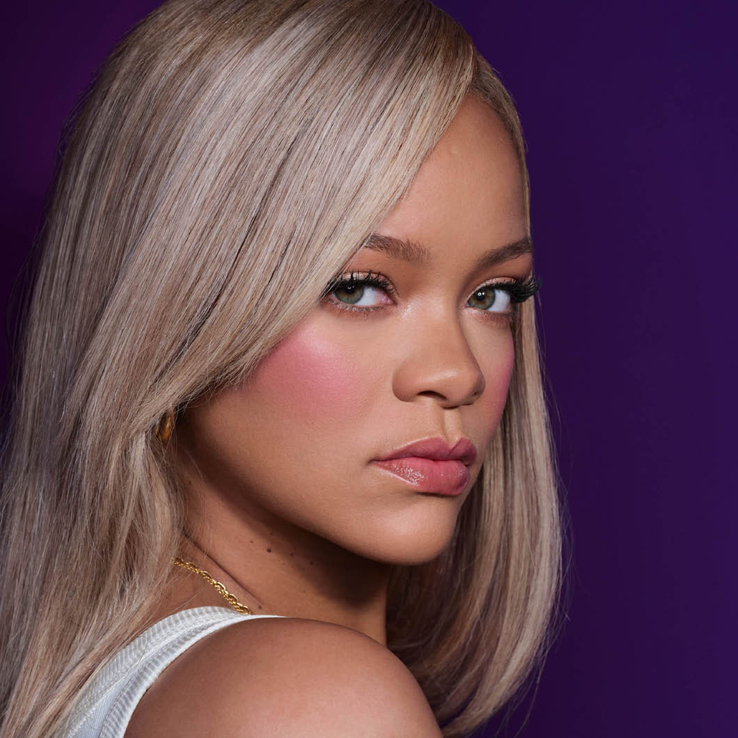 Lähikuva henkilöstä, jolla on vaalea blondi tukka, kiiltävät vaaleanpunaiset huulet, poskipuna ja korostetut ripset tummanviolettia taustaa vasten. Beautylook, jossa korostuvat huulimeikki, silmämeikki ja hehkuva ihomeikki.