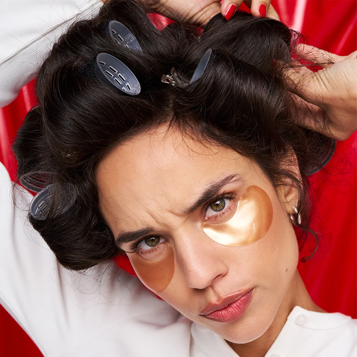 Kvinne med mørkt hår i hårruller og gullfargede øyemasker under øynene står foran en rød bakgrunn. Hun holder hendene i håret og ser konsentrert mot kameraet.