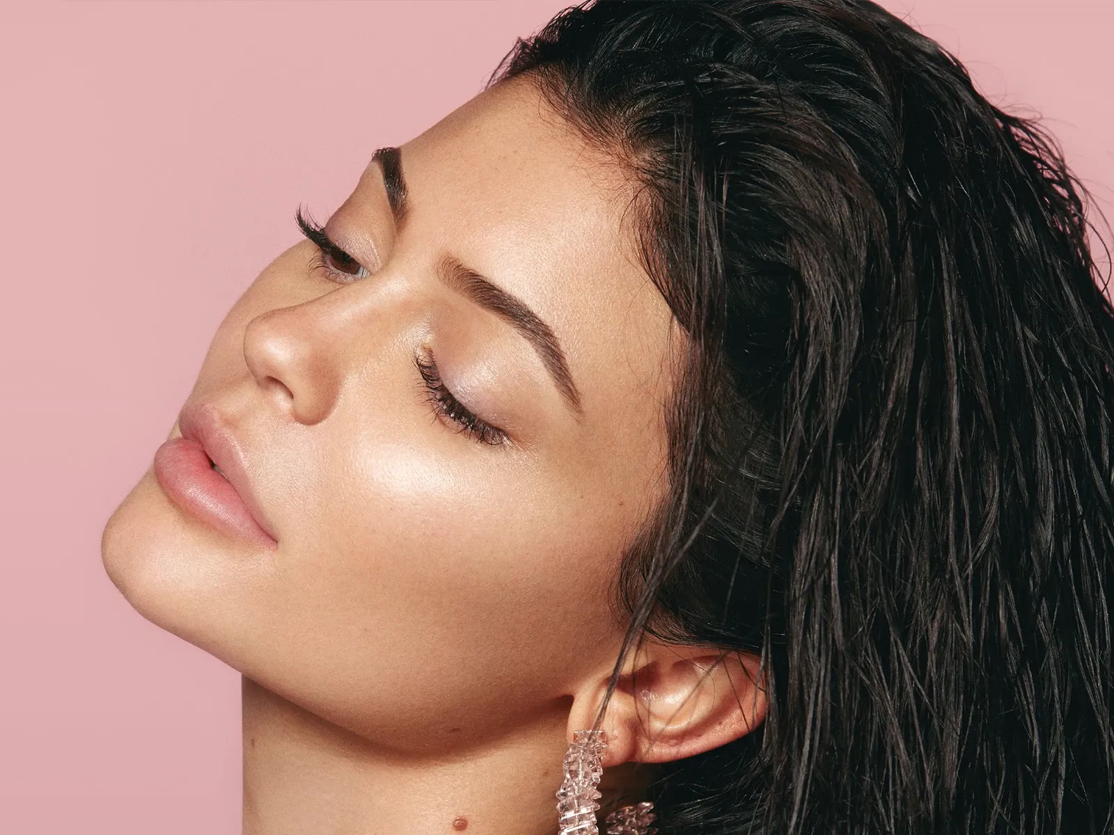 Kylie Jenner i profil som ser ned og lener hodet bakover mot en rosa bakgrunn.