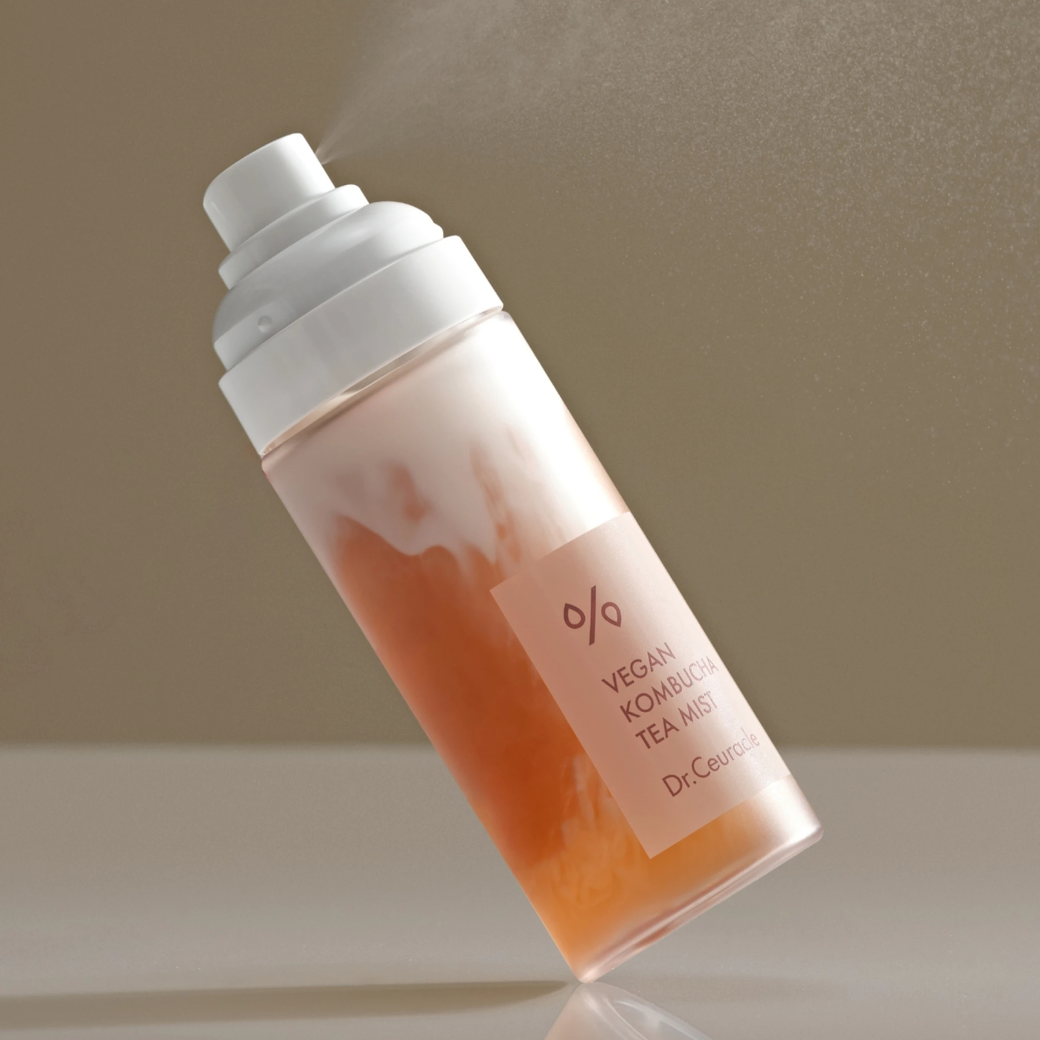 Ansiktsmist i sprayflaska med texten “VEGAN KOMBUCHA TEA MIST Dr. Ceuracle”. En fin dusch av ansiktsmist sprids från flaskan med tvåfasig vätska i beige och orange toner.