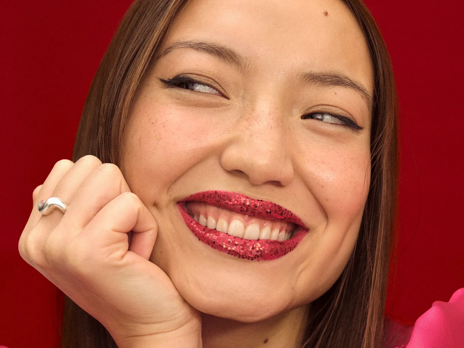 En person med rött glittrande läppstift, rosa tröja, silverring, röd bakgrund.