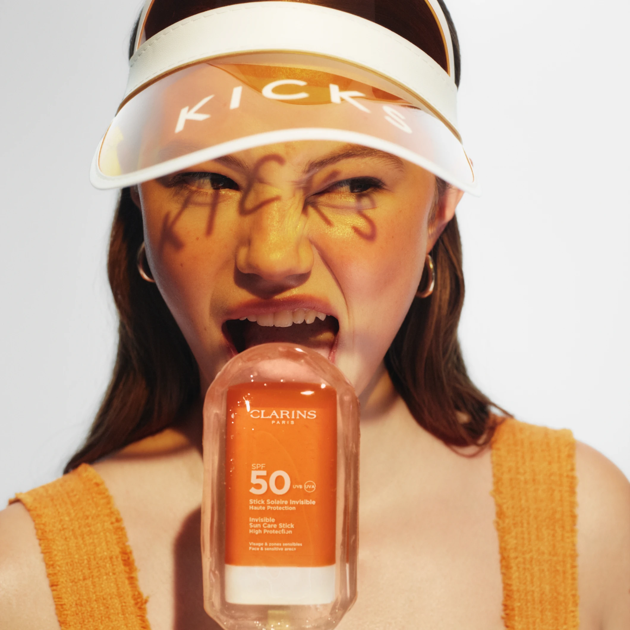Henkilö, jolla on “KICKS”-lippis ja kasvoissa kirjoitettua tekstiä, pitää kädessään aurinkotuotetta “CLARINS SPF 50 Invisible Sun Care Stick High Protection”.