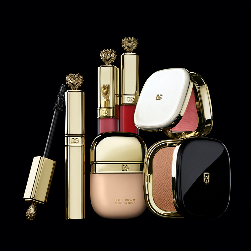 Flere sminkeprodukter fra Dolce & Gabbana, mot en svart bakgrunn. Produktene består av maskara, leppestift, foundation og pudder, alle med elegante gullforpakninger og dekorative detaljer. Leppestiftene har hjerteformede topper, og pudderpakkene har både hvite og svarte lokk med Dolce & Gabbana-logoen. Produktene er ordnet på en måte som understreker den luksuriøse designen.