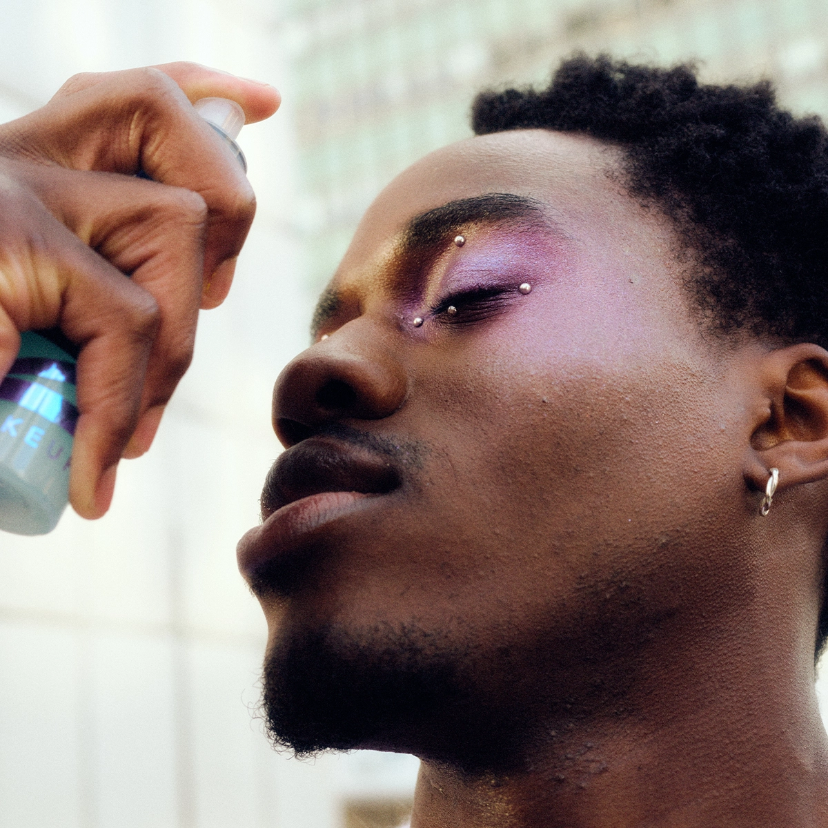 Person med kort hår och färgstark ögonskugga i lila och gult sprayar settingspray mot ansiktet. Närbild i profil med stängda ögon, två piercingar i ögonbrynet och örhänge i örat. Flaska med ansiktsspray hålls nära huden mot ljus bakgrund.