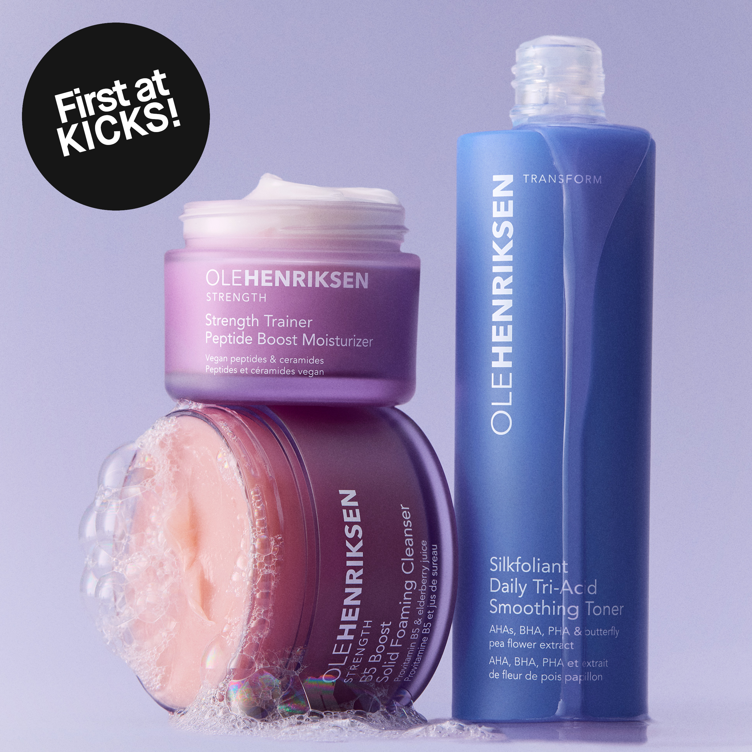 OLEHENRIKSEN Strength Trainer Peptide Boost Moisturizer, Truth Juice Daily Cleanser og Transform Silkfoliant Daily Tri-Acid Smoothing Toner vises mot en lys lilla bakgrunn. En svart sirkel med teksten «First at KICKS!» vises sammen med produktnavn og ingrediensteksten «AHAs, BHA, PHA & butterfly pea flower extract».