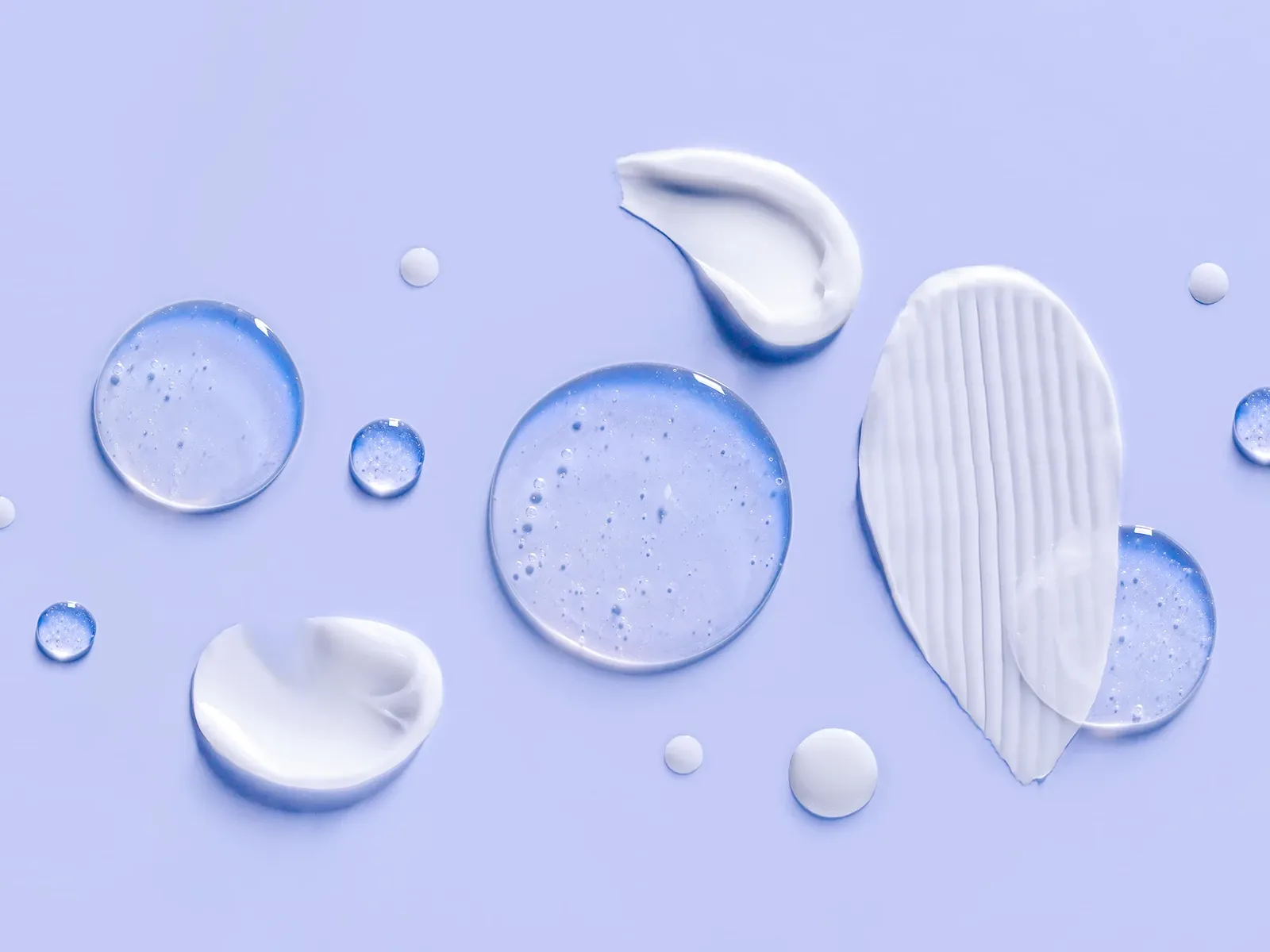Ulike teksturer av hudpleieprodukter – kremer, geler og serum – spredt utover en lys lilla bakgrunn.