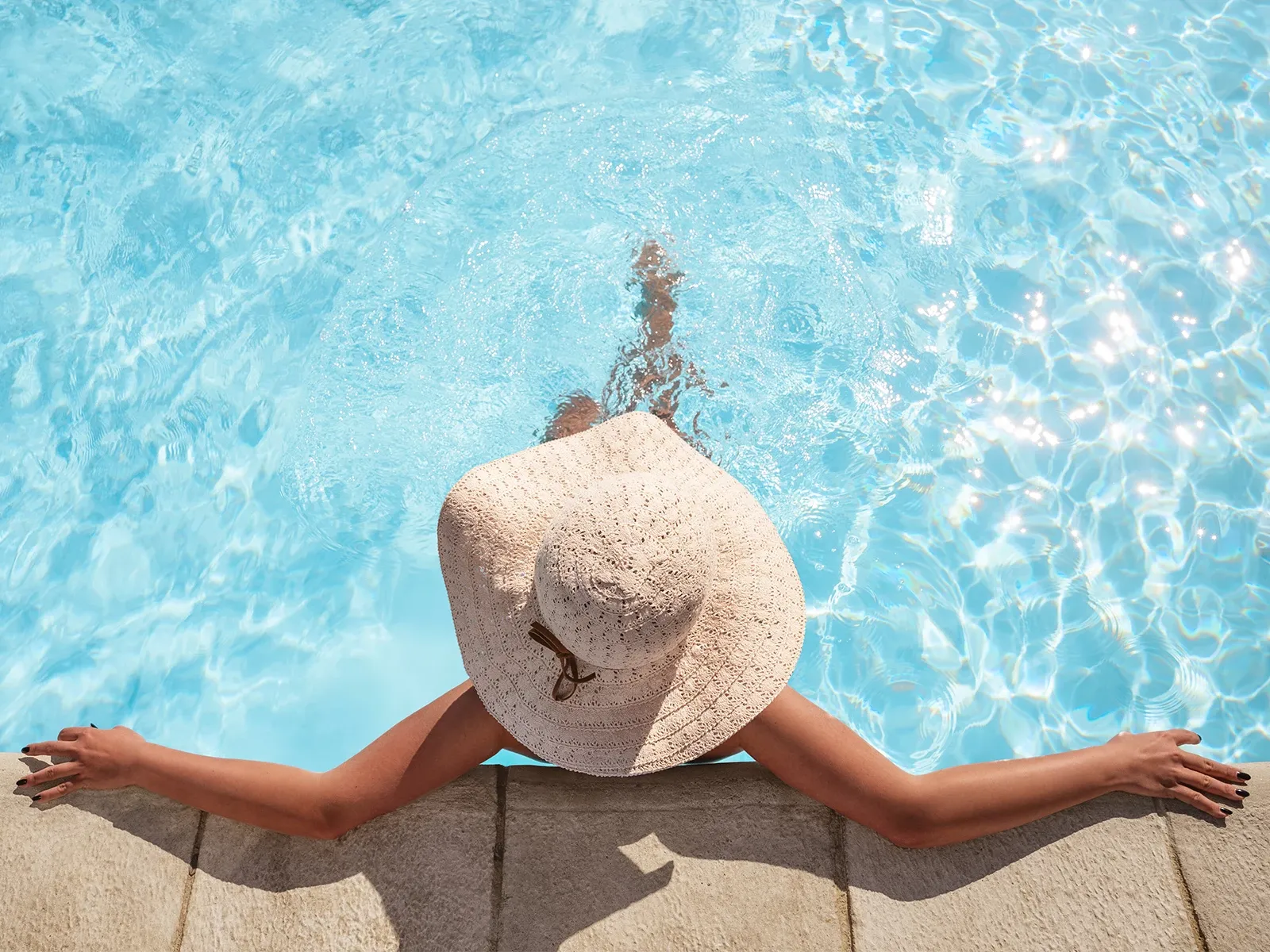 Kvinna med solhatt sitter vid poolkanten med benen i vattn