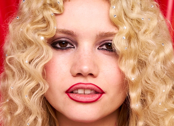 Person med långt lockigt blont hår. Läpparnas konturer målade med röd läppenna. Bakgrunden är röd. 