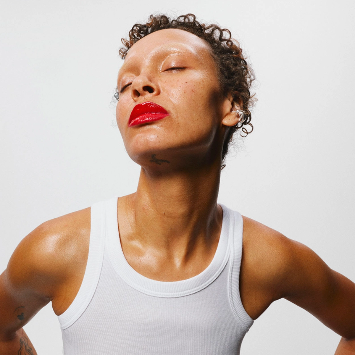 Porträtt i halvprofil av en person mot ljus bakgrund. Personen har kort lockigt hår, slutna ögon och bär rött läppstift. Axlar och övre bröst syns, och personen har ett vitt linne.