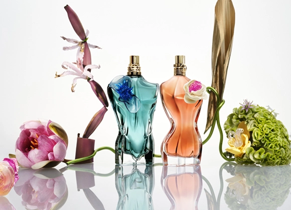 Två Jean Paul Gaultier parfymflaskor, La Belle och Le Beau Flower Edition, i form av kroppar omgivna av färgstarka blommor på en spegelblank yta.