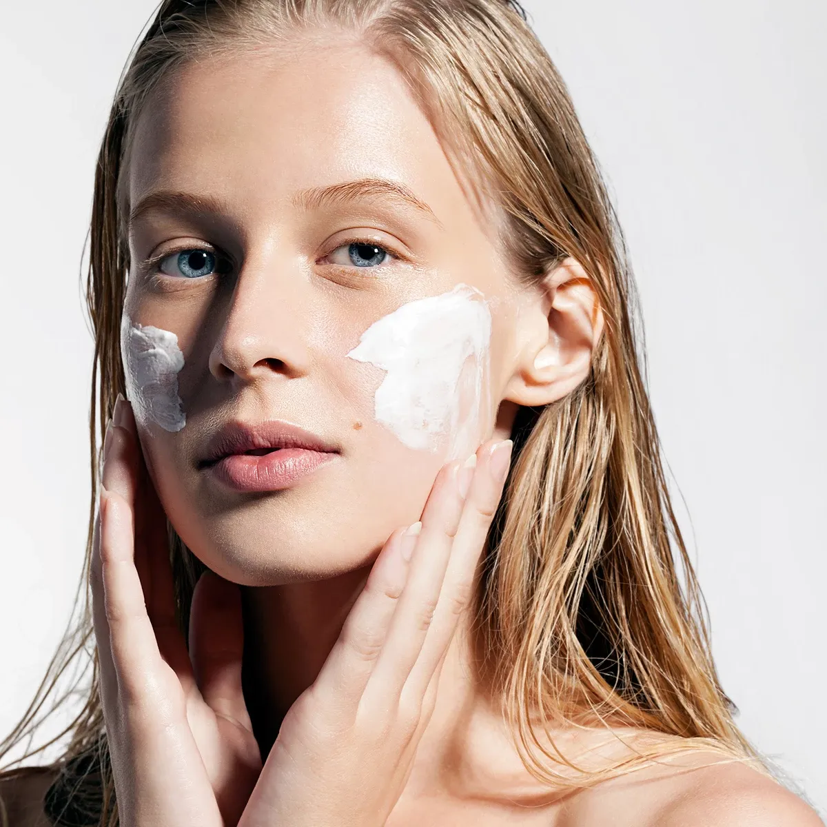 En person med blont hår och händerna vid ansiktet, med vit creme på kinderna
