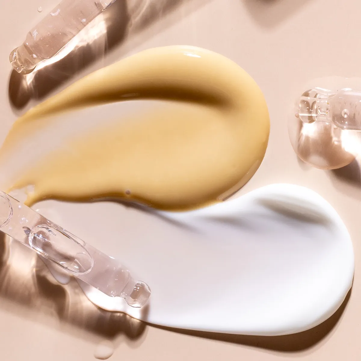 Nærbilde av to forskjellige hudpleieprodukter, en gul og en hvit, påført på en beige overflate med pipetter.
