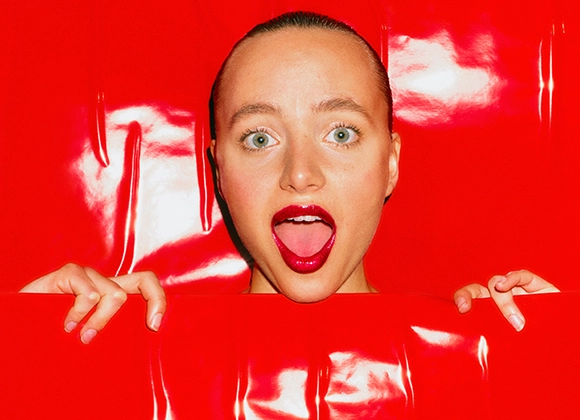 Hodet til en person med gapende munn og røde lepper i midten av et rødt glanset papir, hendene stikker opp.